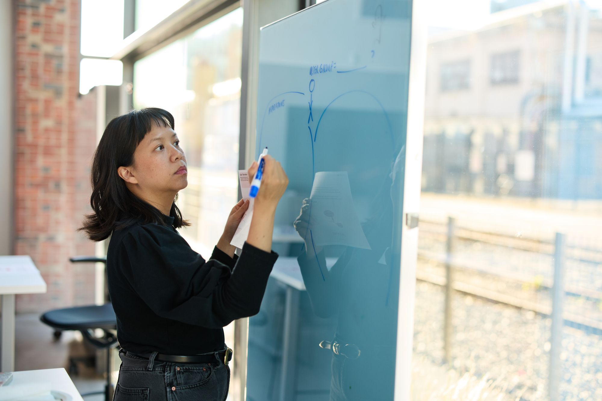 Against the bright full-length windows, a participant thoughtfully writes on the whiteboard.
